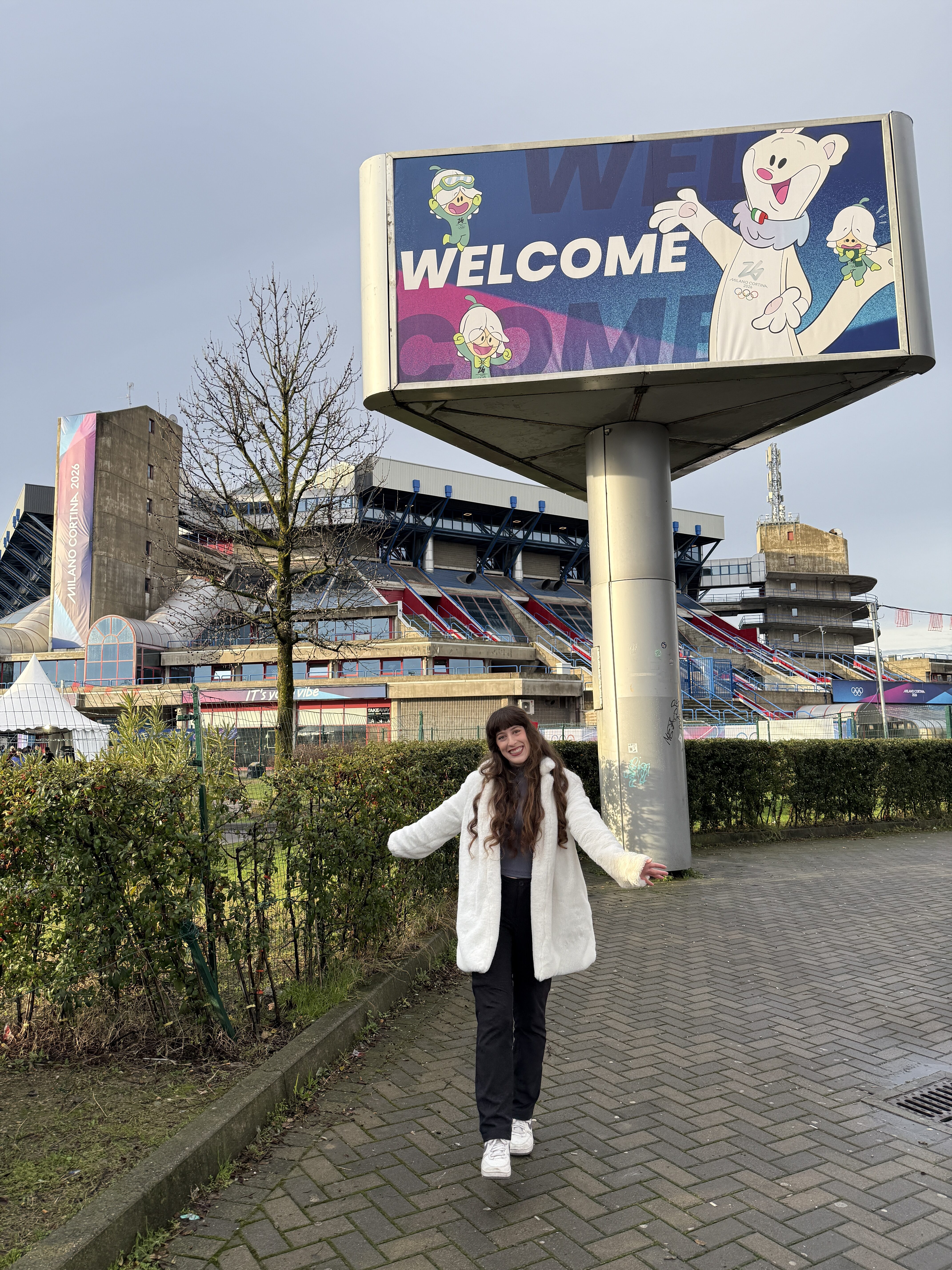 Bella smiling in a fluffy white coat in front of a large WELCOME sign at the figure skating arena (2026 Milano-Cortina)