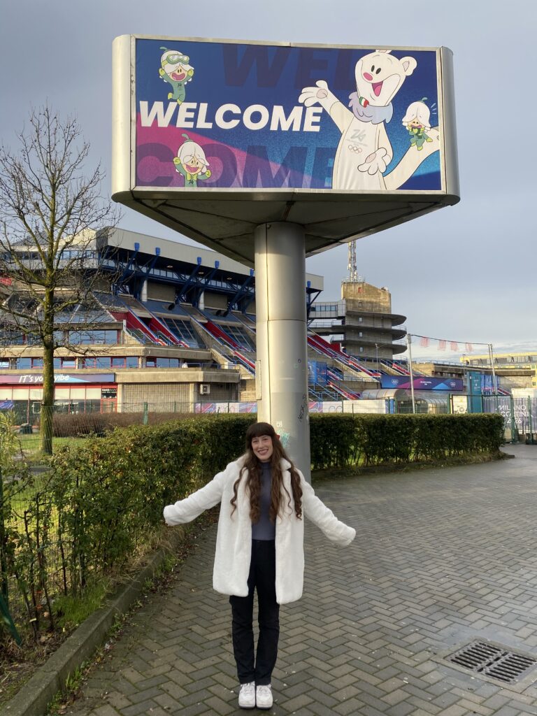 Bella posing in a fluffy white coat in front of a large WELCOME sign at the figure skating arena (2026 Milano-Cortina Olympics)