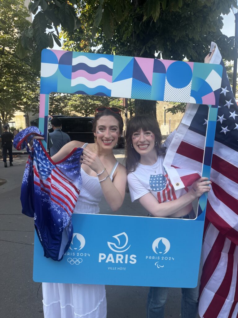 Twin sisters posing with a Paris Olympics photo frame holding leotards and an American flag.