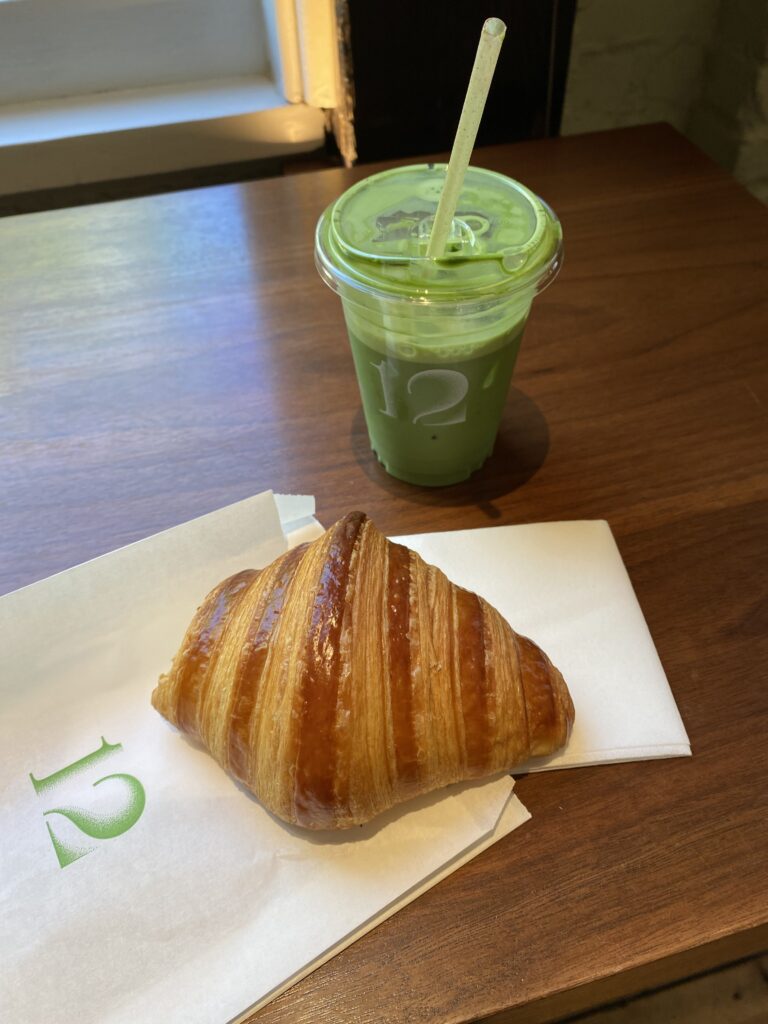 A flaky croissant and a vibrant green matcha on a wooden table with a napkin labeled with a green "12" from 12 Matcha in NYC.