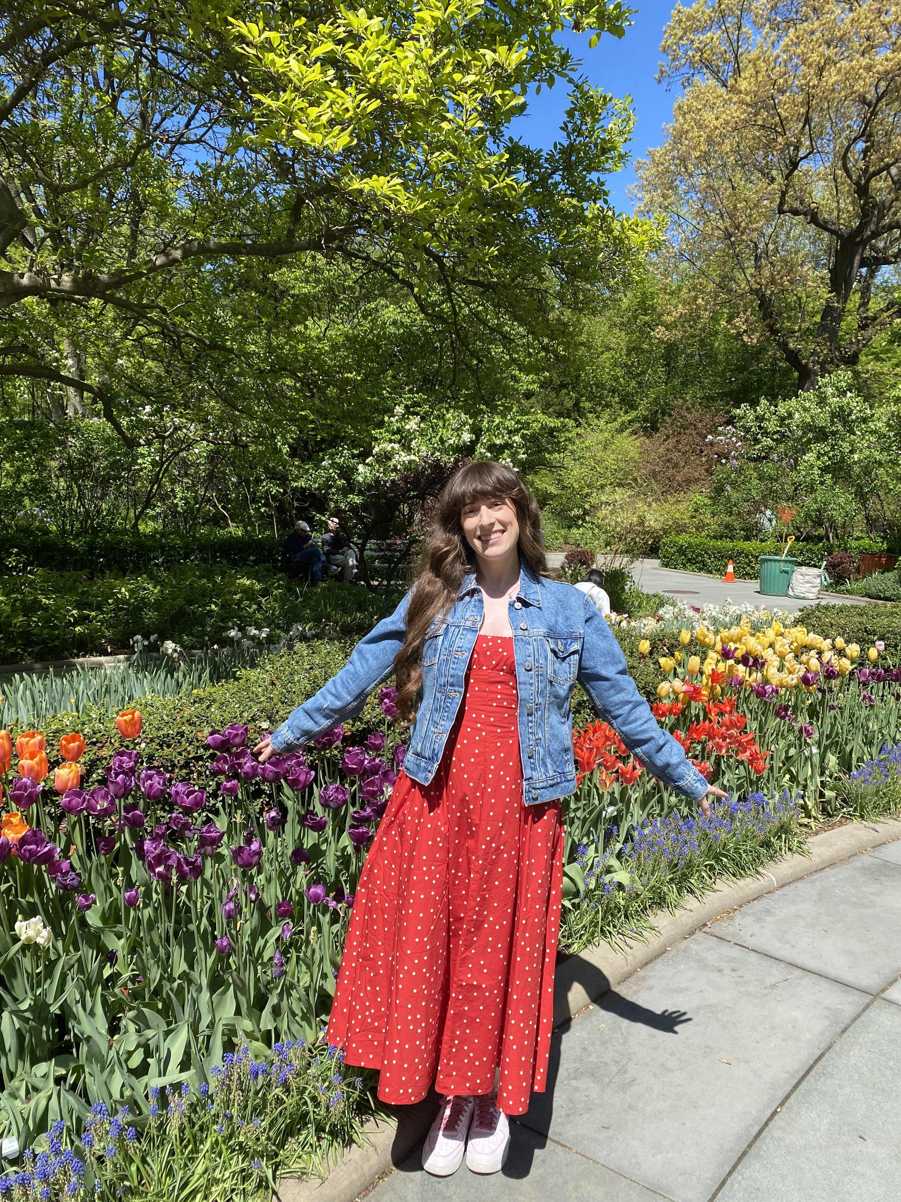 Bella in a red polka dot dress and a jean jacket smiling in front of colorful tulips at the Central Park Conservatory Garden.