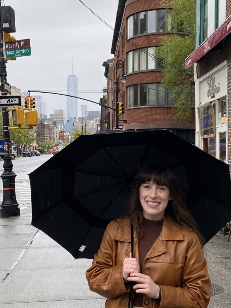 Bella in a leather coat holding a black umbrella on a street NYC's West Village with One World Trade Center in the background.