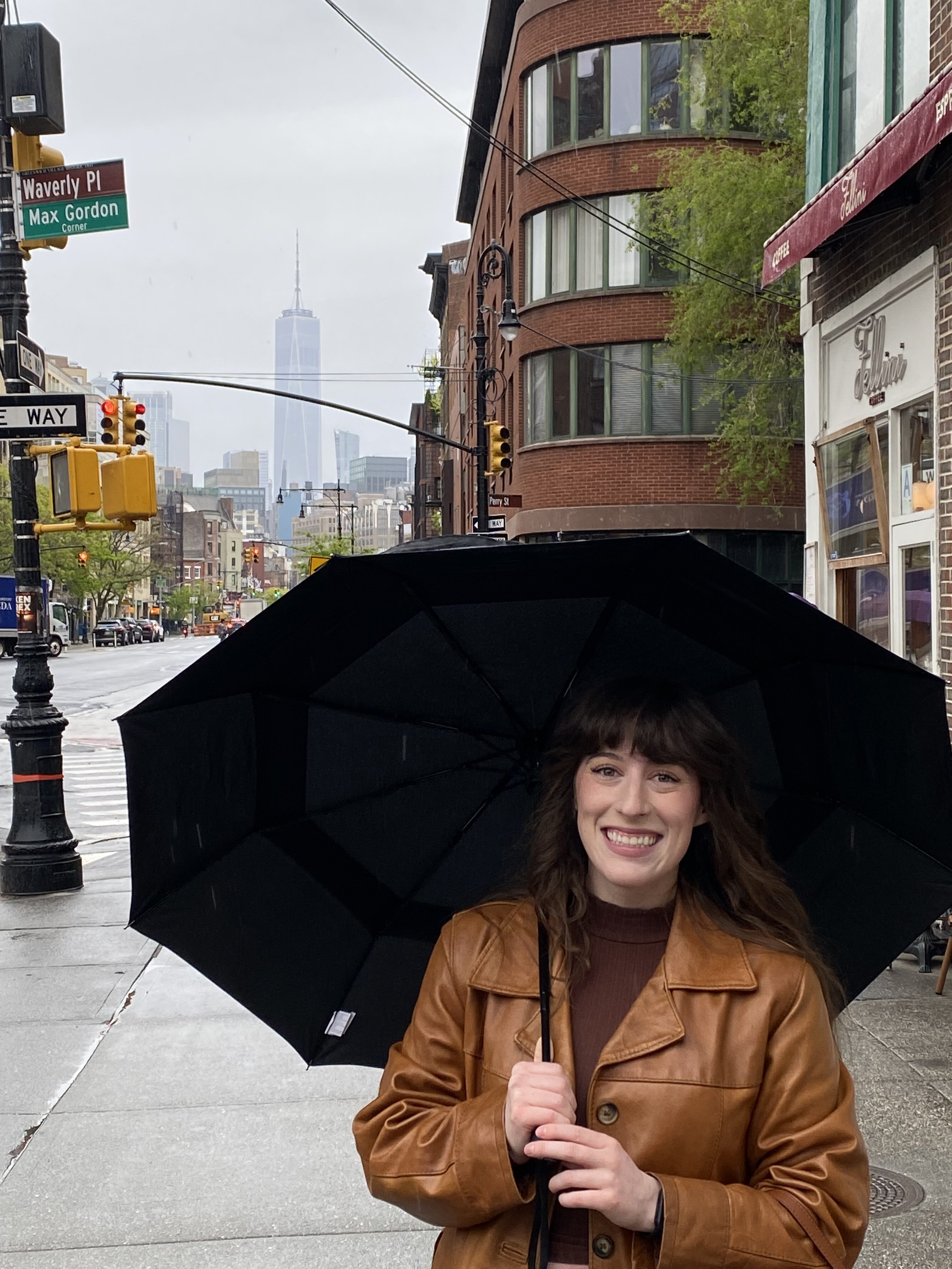 Bella in a leather coat holding a black umbrella on a street NYC's West Village with One World Trade Center in the background.