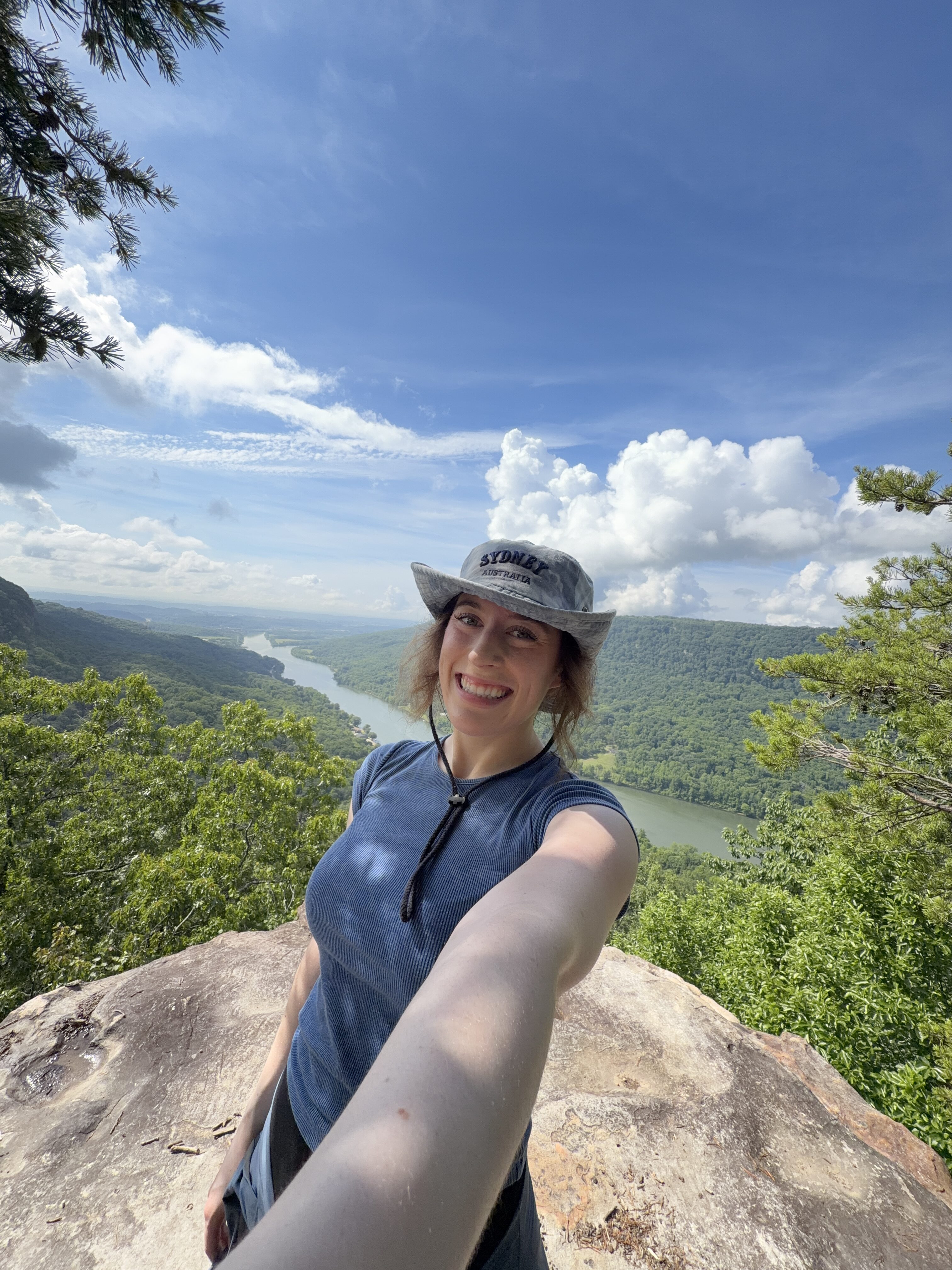 Bella smiling in a blue shirt and a blue hat in front of the Tennessee River Gorge at Edward's Point.