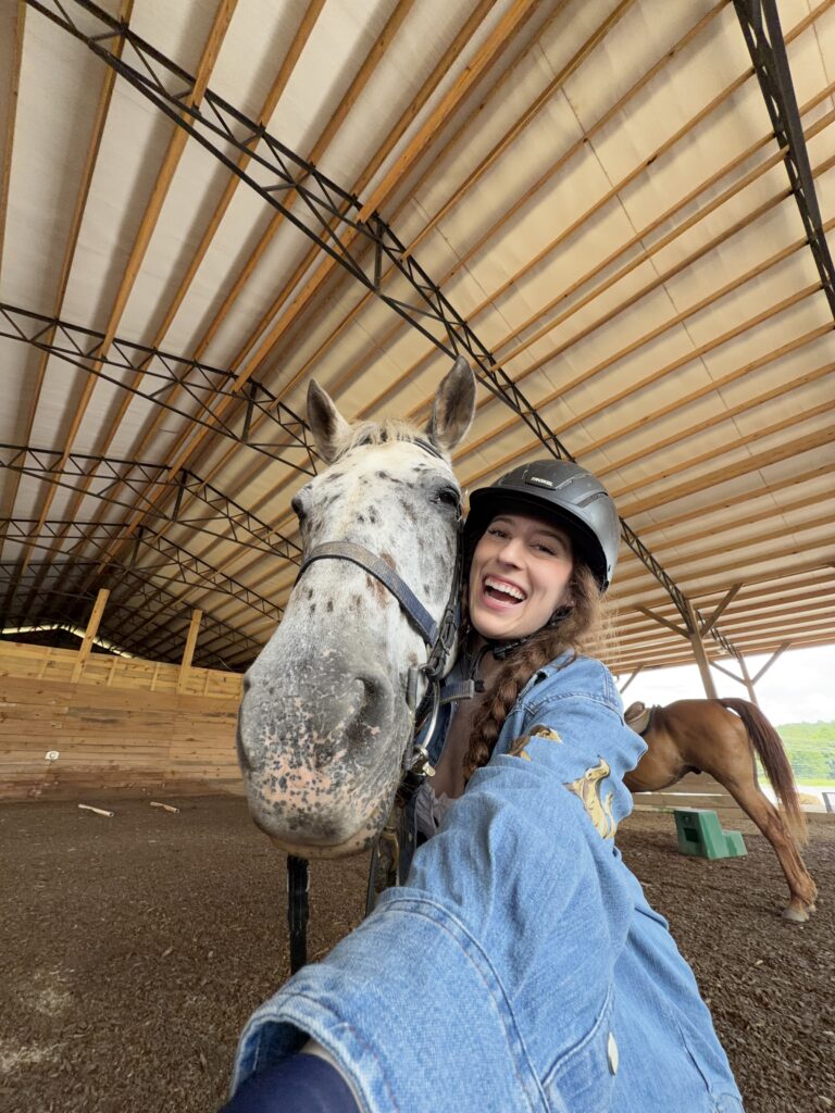 Bella, smiling for a selfie with a grey spotted horse in a covered arena.