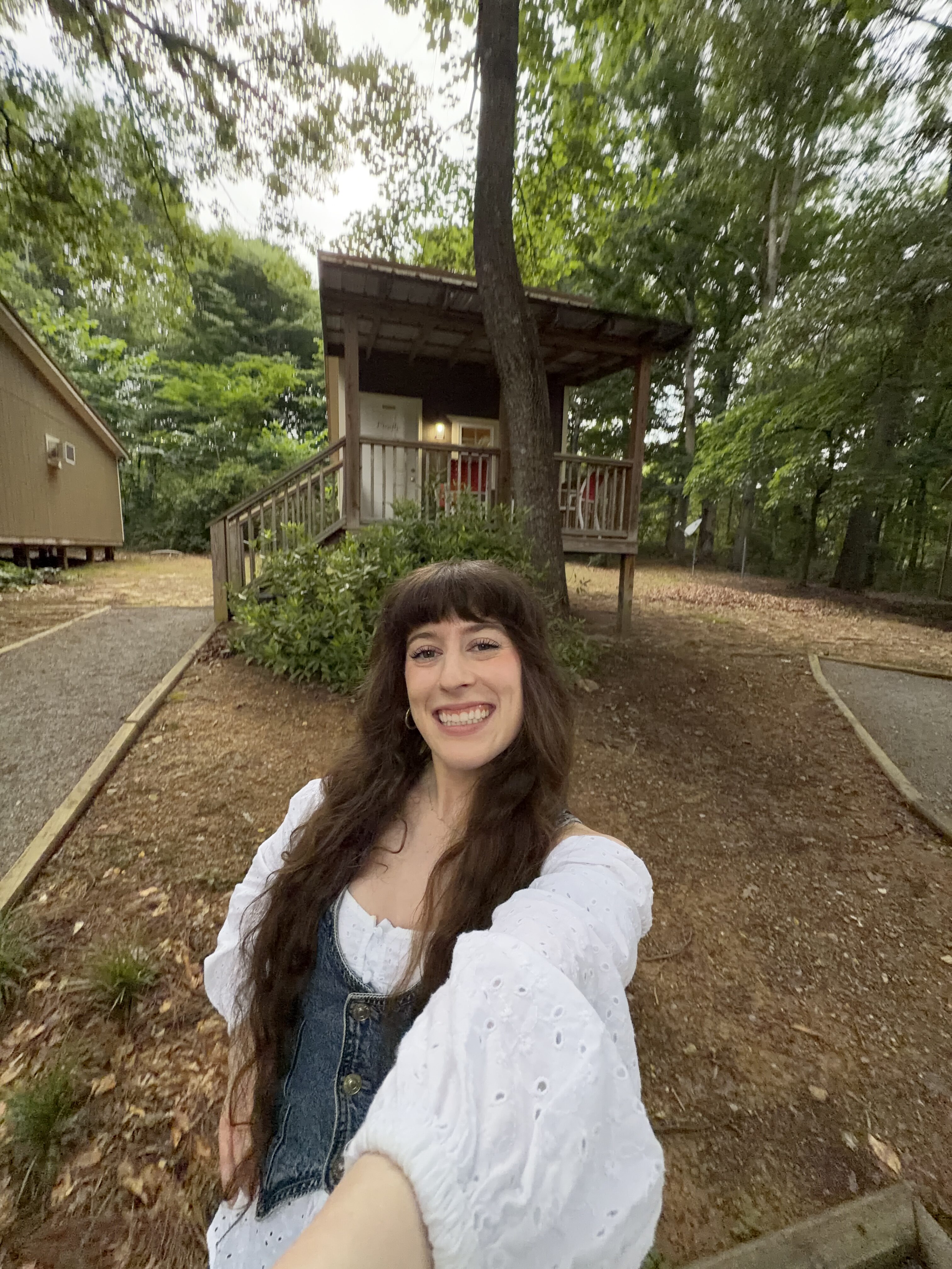 Bella smiling in a flowy white shirt with a jean vest in front of a cozy bungalow cabin at Adventures Unlimited
