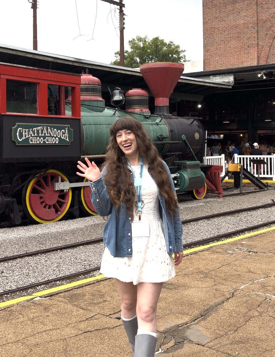 Bella waving in a short white dress with a denim jacket in front of the red, green and black Chattanooga Choo Choo.