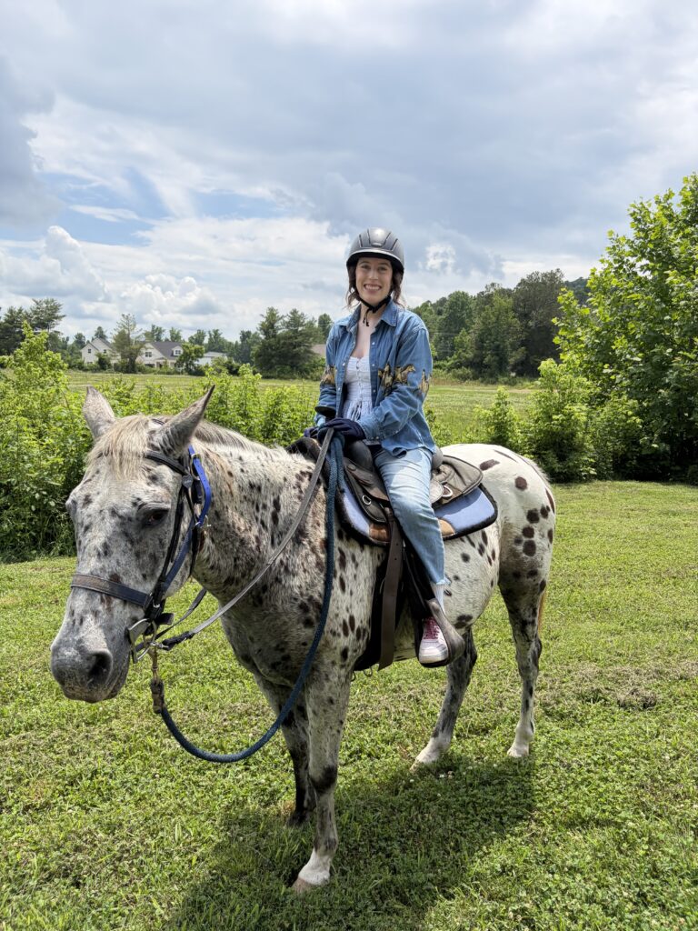 Bella, smiling on a grey and white spotted horse among grassy fields and trees, Hidden Hills Farm.