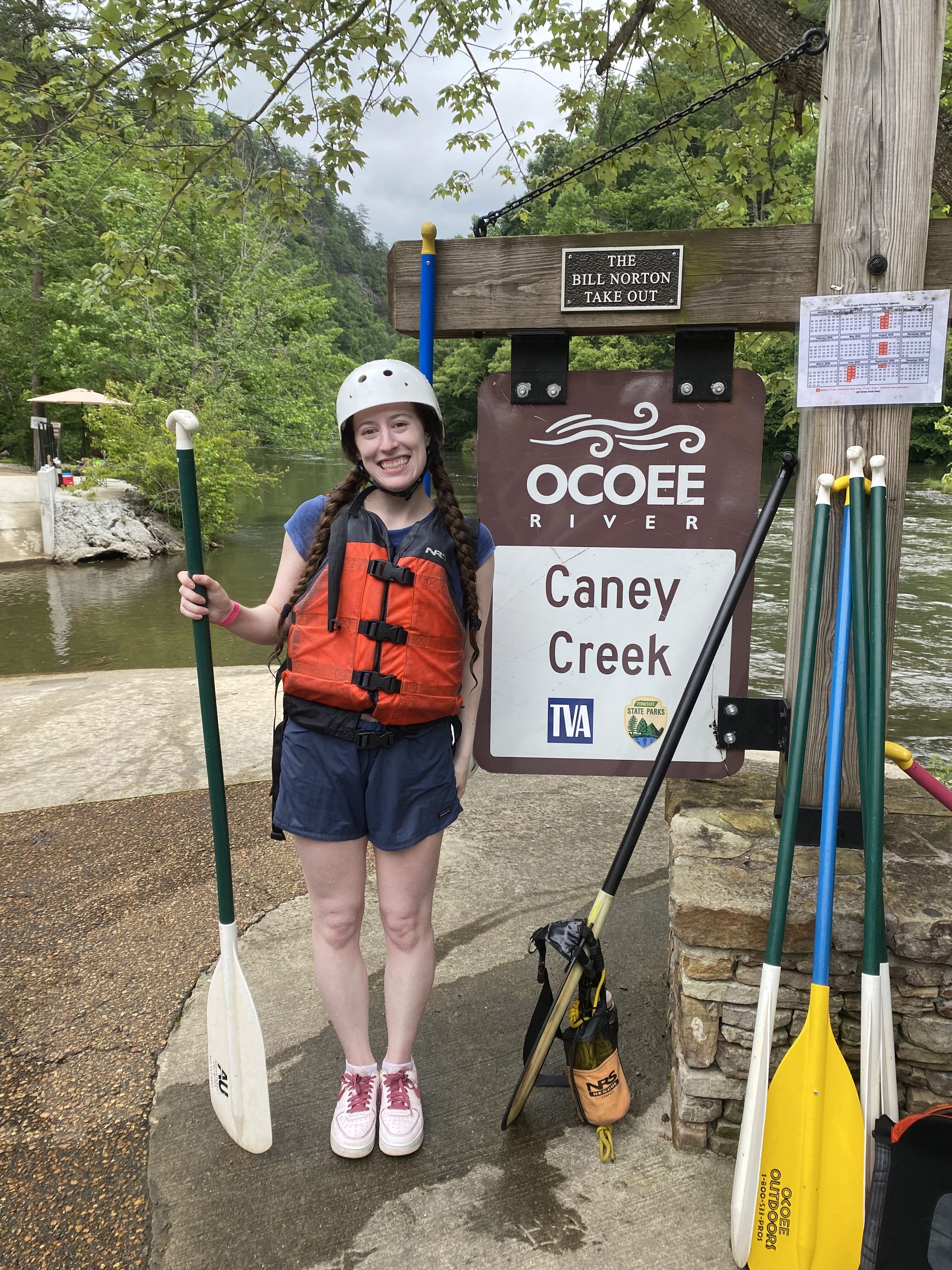 A young woman with blue shorts and an orange life vest posing with her rafting paddle in front of the Ocoee Caney Creek sign.