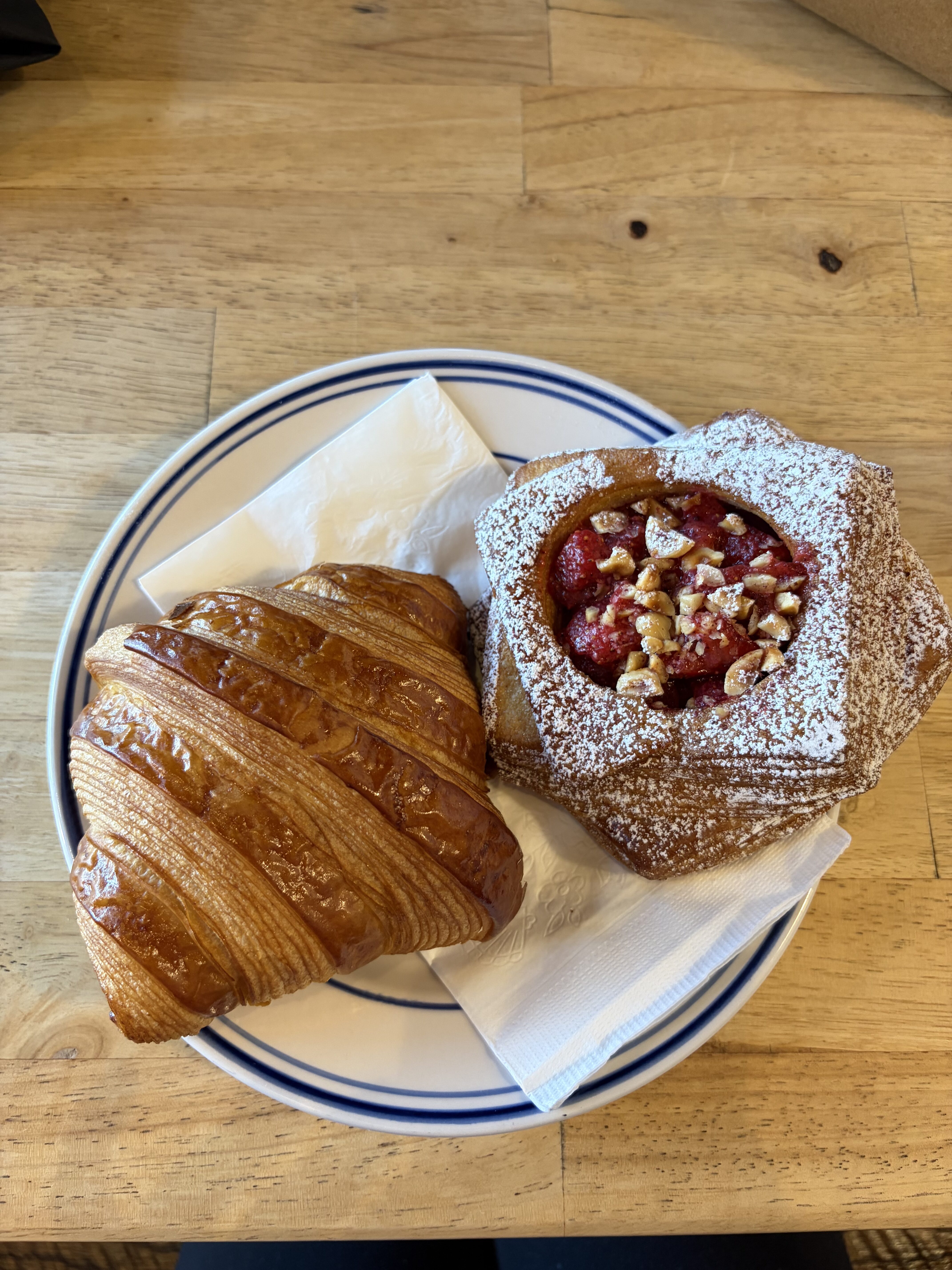 A small plate with a plain croissant and a powdered sugar-dusted pastry with fruit filling, on a wooden table at Niedlov's.