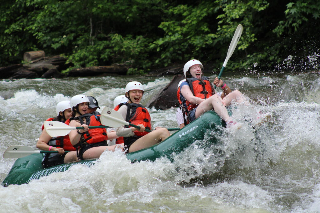 A group of river rafters navigating a tumultuous rapid, while a daring young woman sits on the bow (front) of the raft.