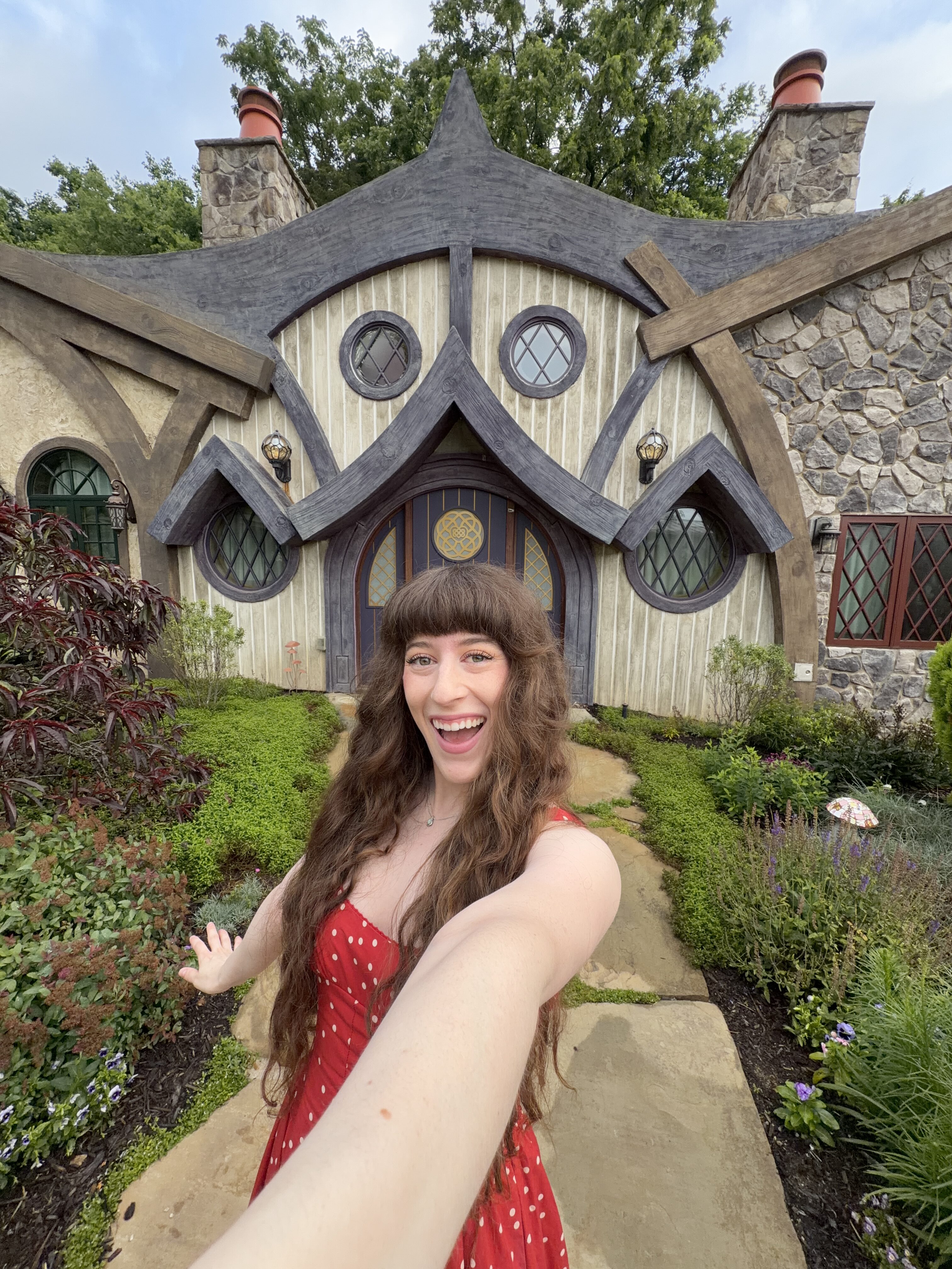 A young woman with brunette hair smiling in a selfie in her red dress in front of the purple-outlined fairy cottage at the Ancient Lore Village. 