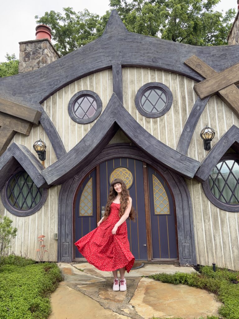 A young woman posing with her red dress in front of a cottage with plum trim and weathered grey wood at the Ancient Lore Village.