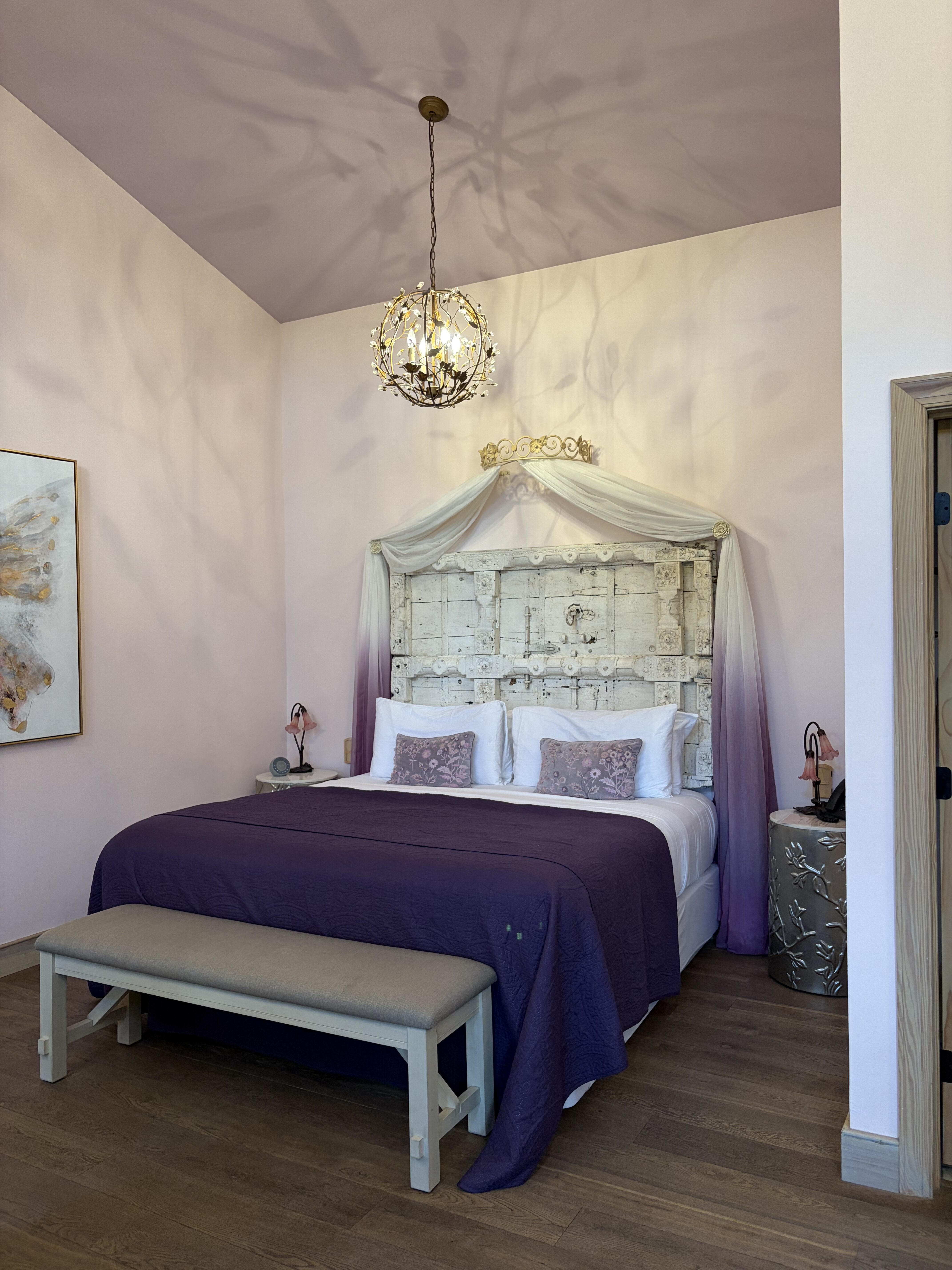 A King bed with dark purple bedding, light purple pillows and a wooden bed frame, surrounded by dainty pink walls at the Ancient Lore Village.