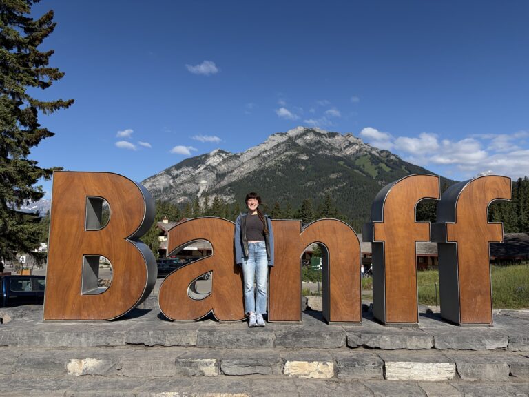 Bella smiling in front of the "Banff" sign, squinting in the sun.