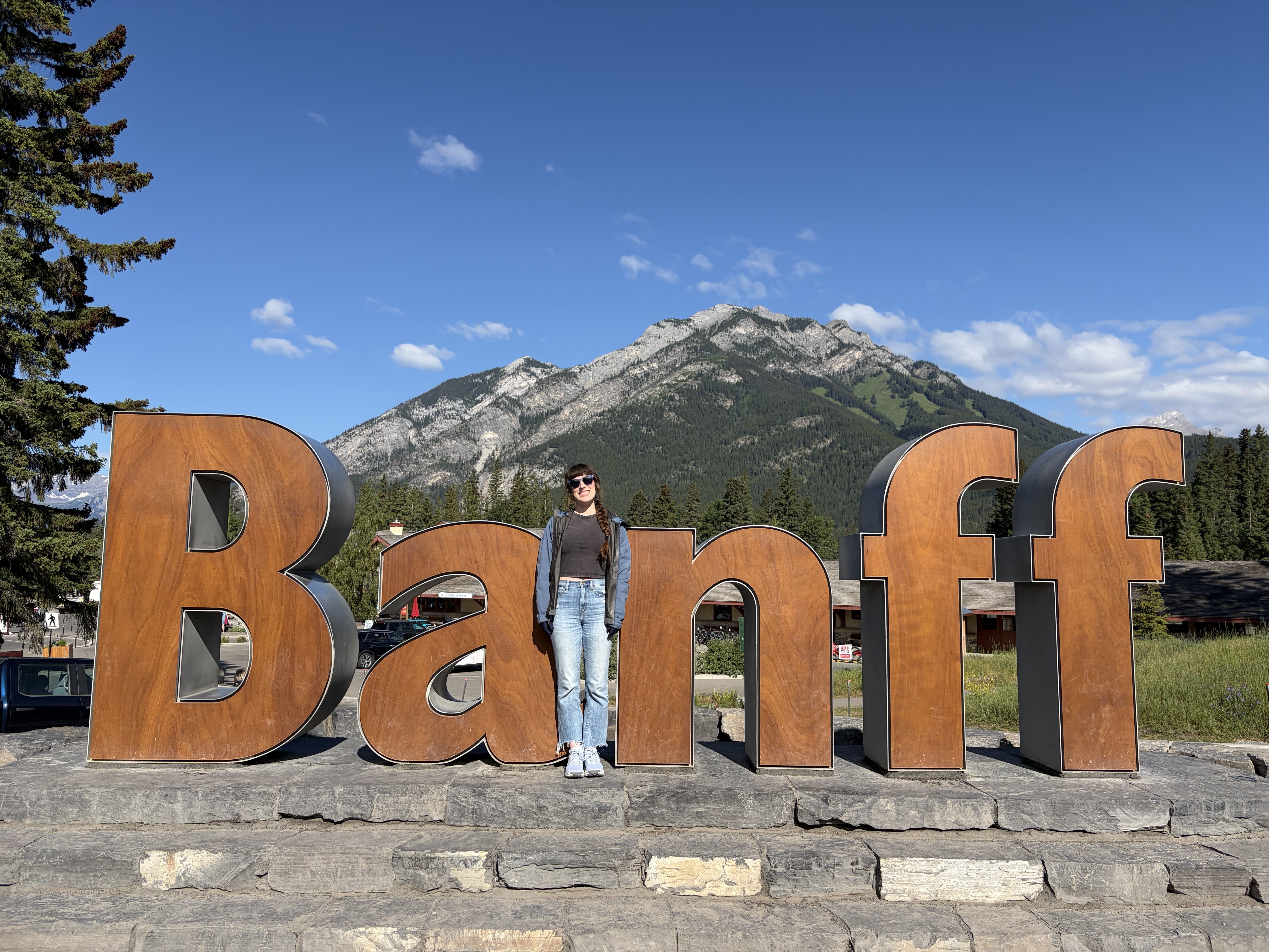 A woman standing in front of the wooden "Banff" sign with Mt. Norquay in the background. 