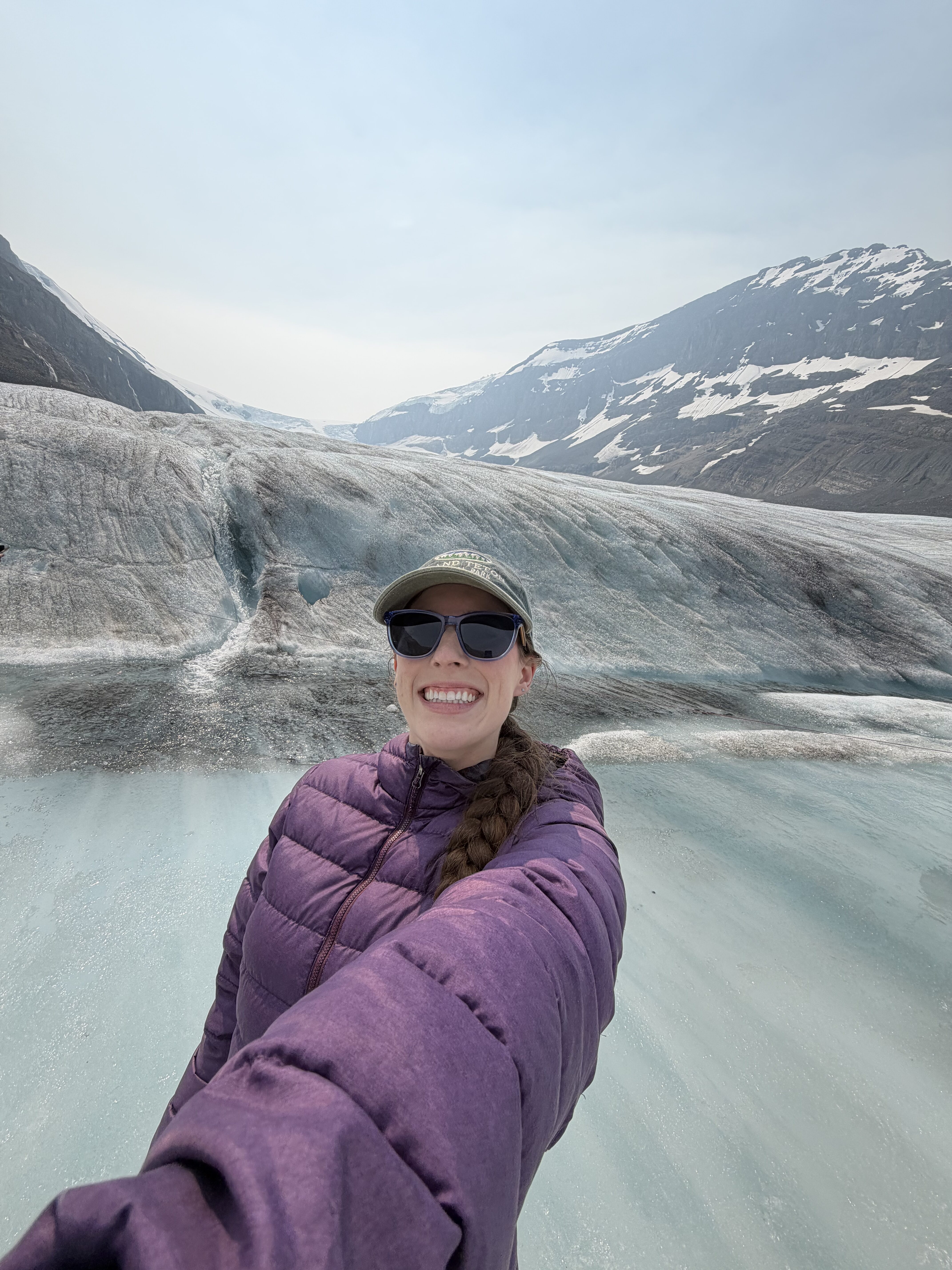 Bella, wearing a purple coat, sunglasses and a hat, smiling (selfie) from on the Athabasca Glacier.  