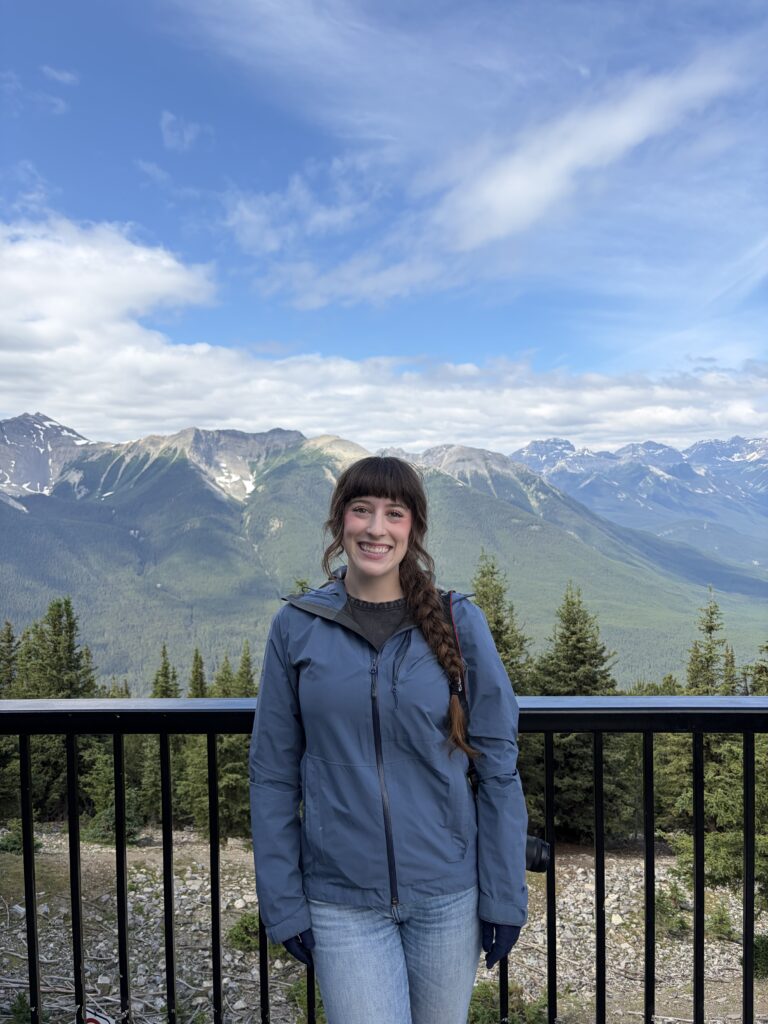 A young woman wearing a blue raincoat smiling at the Banff Gondola lookout deck with the Canadian Rocky Mountains in the background.