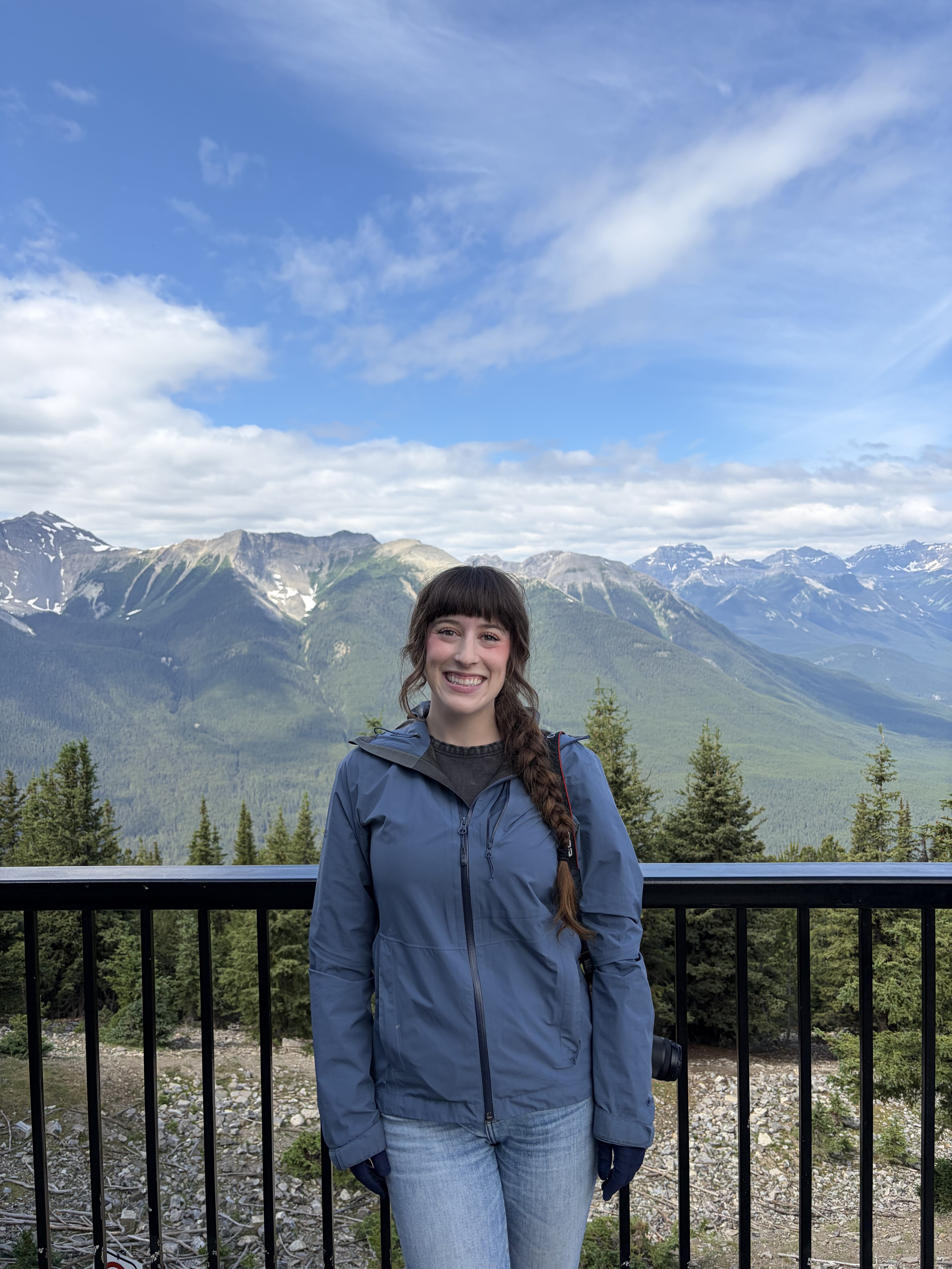 A young woman wearing a blue raincoat smiling at the Banff Gondola lookout deck with the Canadian Rocky Mountains in the background. 