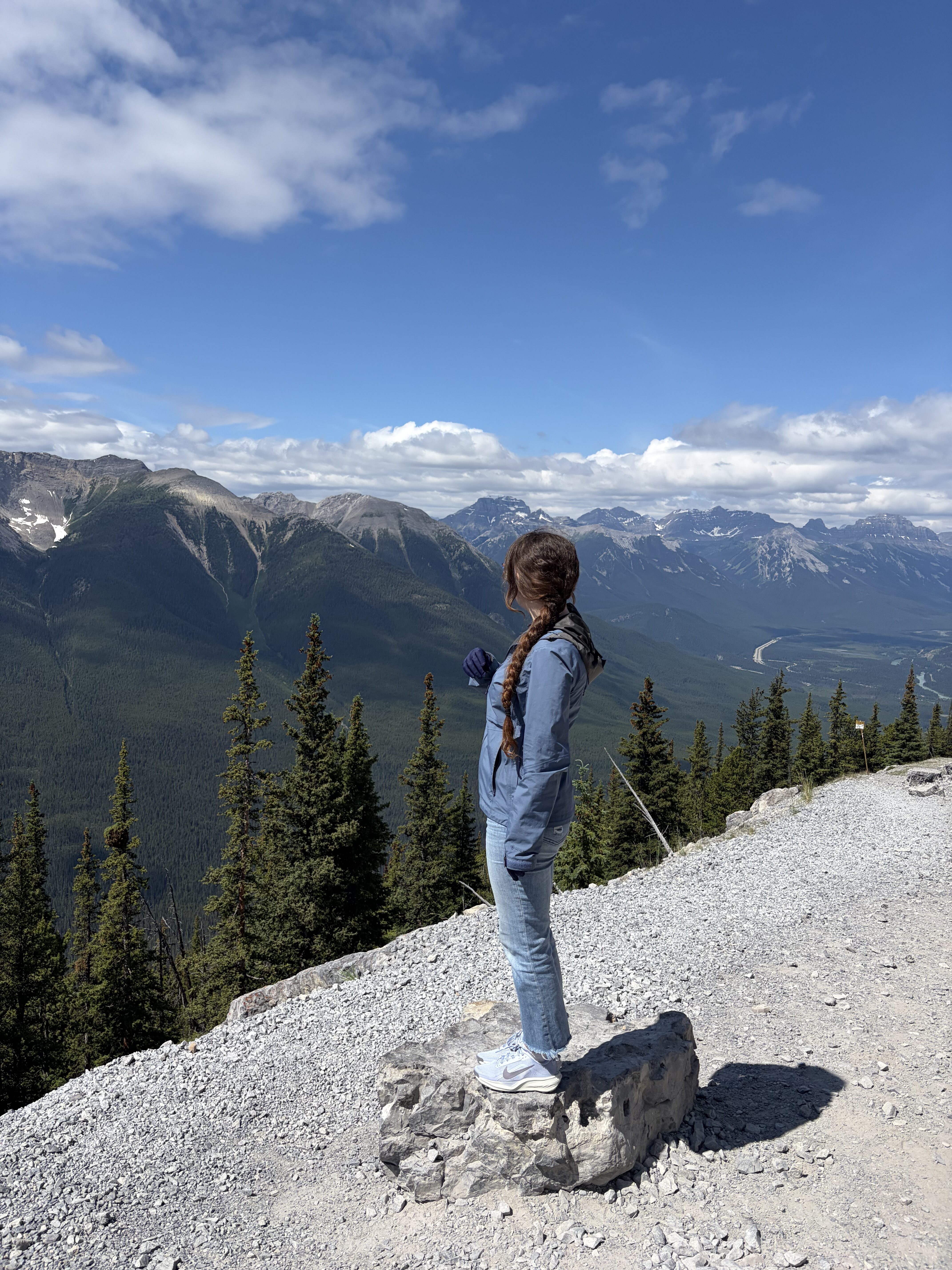 A young woman standing on a grey rock situated on a grey gravel trail, gazing out at the Canadian Rocky Mountains at the top of Banff Gondola.